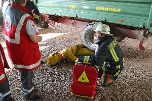 Bild: Ein Arbeiter ist verletzt und unter einem Anhänger eingeklemmt. Helfer des DRK und Einsatzkräfte der Feuerwehr übernehmen die Versorgung und Befreiung.